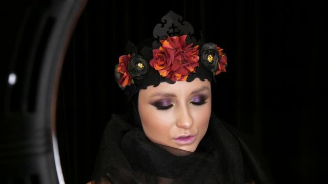 Professional girl model with beautiful makeup poses in a black cap and wreath on her head in front of the camera on a black background in the image of a black widow. High-fashion