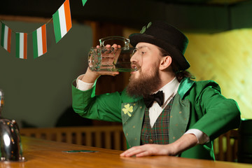 Dark-haired bearded young man in a leprechaun costume drinking green beer from a big glass