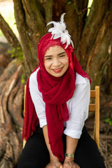 Women muslim, women islamic,wearing a red fabric head covering adorn with white fabric flowers wear shirt white sit poses photography on the chair under the tree before to ramadan month.