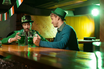 Dark-haired bearded young man in a leprechaun hat and his friend spending evening in the pub