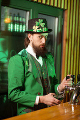 Dark-haired bearded young man in a leprechaun hat pouring green beer into the glass