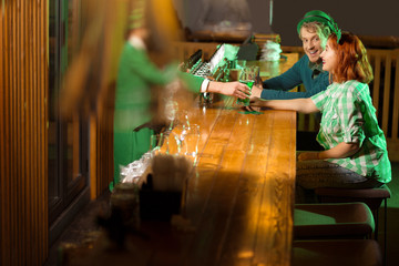 Dark-haired bearded young man in a leprechaun hat giving a glass of beer to a girl