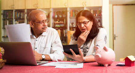 Senior Indian/asian couple accounting, doing home finance and checking bills with laptop, calculator and money while sitting on sofa/couch at home