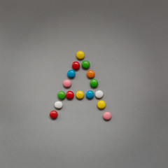 Capital letter A made of multicolored chocolates in the glaze on a gray textural background.