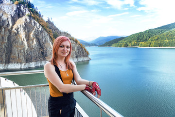 Fototapeta na wymiar girl traveler with red hair posing in background Dam lake Vidraru in Romania, autumn sunny day