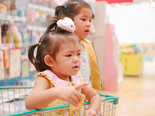 Little Asian baby girl, 24 months old, having fun standing in a shopping cart with her sister seeing many products for sale in a supermarket