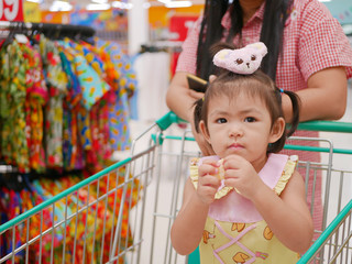Little Asian baby girl, 24 months old, eating snack and standing in a shopping cart while her mother does a shopping in a supermarket / mall