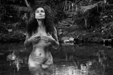 A young  brunette female with a curly hair stands in the tropical creek. Nu style (Naked).Black and White photography. Image. 