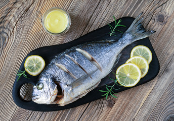 sparus aurata baked in oven on a black serving board decorated with lemon and rosemary with a glass jar of hollandaise sauce on wooden surface 