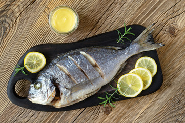 gilt-head bream baked in oven on a black serving board decorated with lemon and rosemary with a glass jar of hollandaise sauce on wooden surface