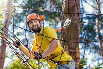 Young male man adult wears protective helmet with action camera having fun in extreme rope park, amusement park. Climbing in rope bridge at green forest. Active healthy lifestyle in spring or summer.