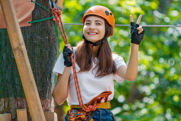 Young beauty female woman adult wears protective helmet having fun in extreme rope park, amusement park. Sitting in rope bridge at green nature forest. Active healthy lifestyle in spring or summer.