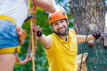 Woman and man in protective gear are walking at adventure park with obstacles and ziplines. Instructor is helping girl to climb rope trail on high tree. Extreme rest and summer activities concept.