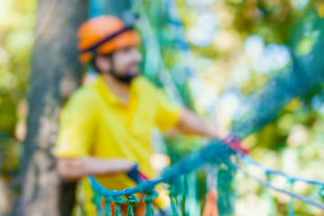 Obraz na płótnie Canvas Young male man adult wears protective helmet with action camera having fun in extreme rope park, amusement park. Climbing in rope bridge at green forest. Active healthy lifestyle in summer. Bokeh