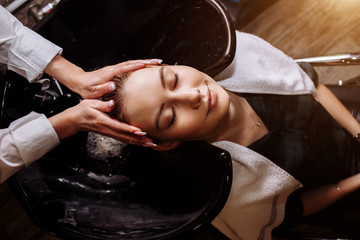 Gorgeous cute young woman enjoying head massage while professional hairdresser applying shampoo her hair. Close up of hairdresser's hands washing hair to the client.