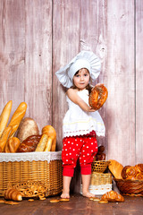 Eat fresh bread and homemade pastries. Little smiling girl in a cook cap with bagel in hand near a wicker basket with bread rolls and bakery products.