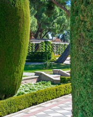 A checkered tiled path comes into view from behind a thick green hedge showing small stairs and a gallery lined with spiral vines wrapped around pillars in the background.