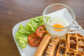 Homemade delicious american breakfast with soft-boiled egg, waffles, sausage, tomato, lettuce on white plate on wood table, top view.