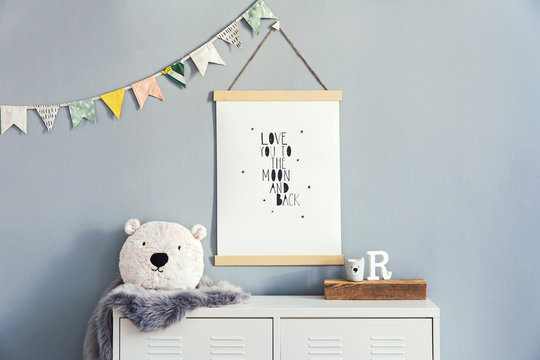 Stylish and cute scandinavian decor of child room with mock up poster, white shelf, natural toys, hanging cotton flags,  accessories and teddy bears. Bright and sunny interior. Grey backgrounds wall.