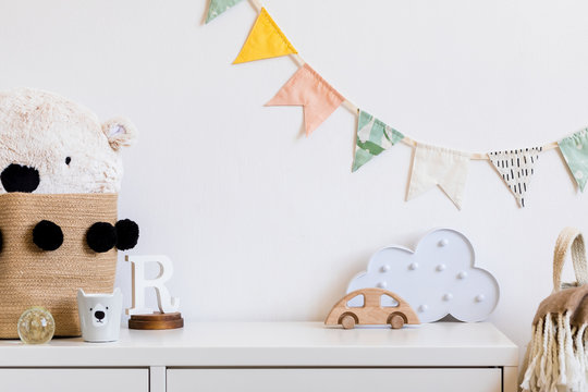Stylish and modern scandinavian child room with hanging cotton colorful flags on the white wall, boxes, teddy bear in natural basket, toys. wooden accessories and cloud. Real photo. 