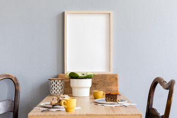 Stylish and retro interior of kitchen space with small wooden table with mock up photo frame, plant, box, design cups, furnitures and tasty dessert. Scandinavian room decor with kitchen accessories.