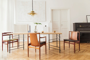 Eclectic and elegant dining room interior with design sharing table, chairs, gold pedant lamp, abstract paintings, piano and stylish accessories. Minimalistic decor. Brown wooden parquet. Real photo.