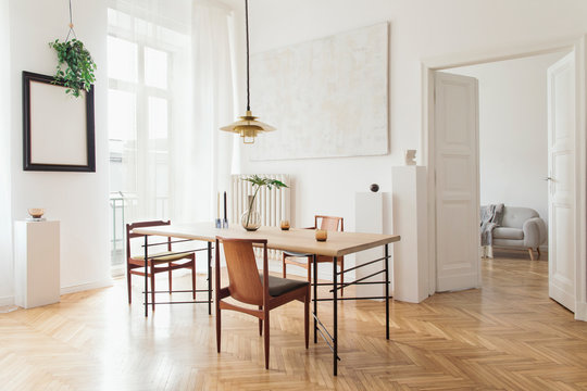 Eclectic and elegant dining room interior with design sharing table, chairs, gold pedant lamp, abstract paintings and stylish accessories. View to living room. Minimalist decor. Brown wooden parquet.