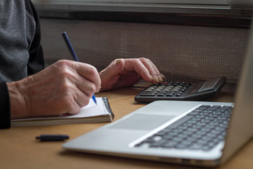 The hands of an elderly man count on a calculator and make notes in a notebook on the background of an open laptop.