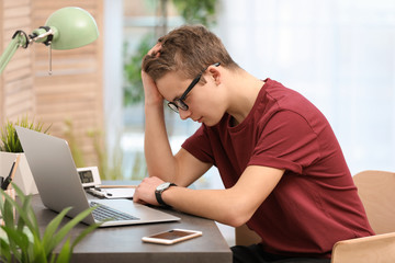 Troubled teenage boy with laptop at table in room