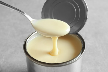 Condensed milk pouring from spoon into tin can on grey background, closeup. Dairy product