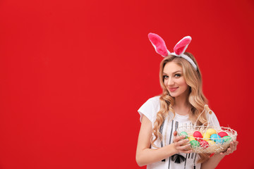 Beautiful young woman in bunny ears headband holding basket with Easter eggs on color background, space for text
