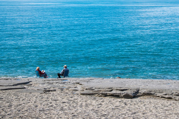 Old pair sitting by the sea, relaxing. Horizontal. With azure sea, send and rocks. Watching the...