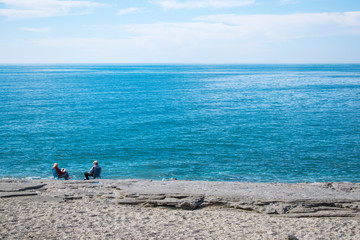 Old pair sitting by the sea, relaxing. Horizontal shot. With sky, azure sea, sand and rocks. Talking.