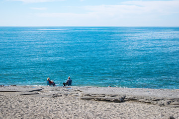 Old pair sitting by the sea, relaxing. Away view. With sky, azure sea, send and rocks. Large copy...