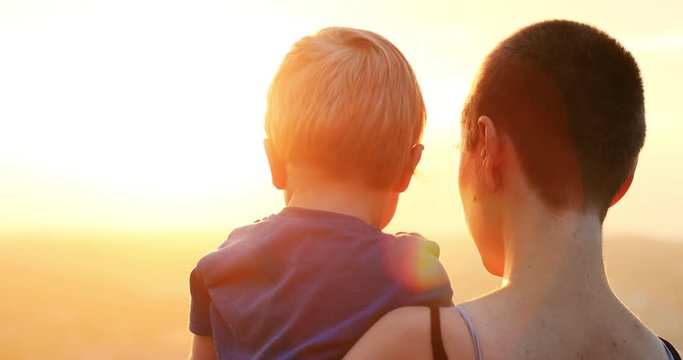 Close-up of beautiful young woman holding her small kid watching amazing sunrise over the city. Back view of mother and son at sunrise.
