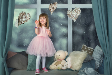 Beautiful little girl in a pink dress and white pantyhose is standing and laughing on a winter evening on the windowsill with fur pillows and soft toys, growth portrait