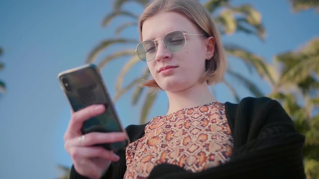 Portrait of young beautiful happy teenager with short blond hair in stylish sunglasses using smartphone to check social networks, mail, Instagram, messenger background palms blue sky slow motion