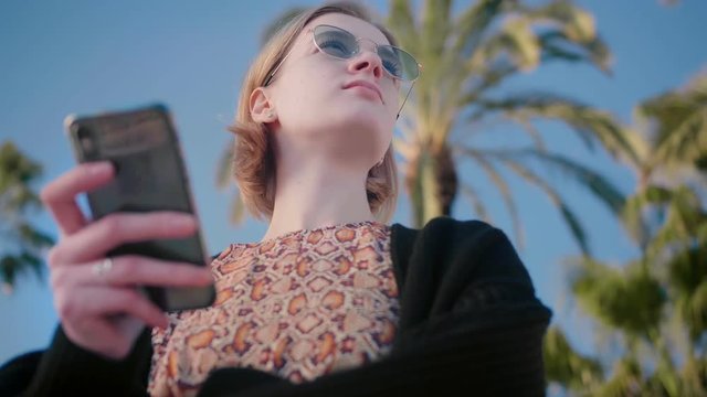 Close up of young beautiful trendy teenager girl in stylish sunglasses waiting for her friends using smartphone to check social networks, mail, Instagram background palms blue sky slow motion
