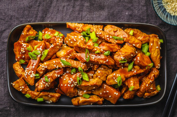 Teriyaki chicken with sesame, green onion in black plate, dark background.