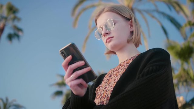 Portrait of young beautiful blond teenager girl in stylish sunglasses using smartphone typing messages, post to social networks, mail, Instagram feeling bored, disappointed background palms blue sky