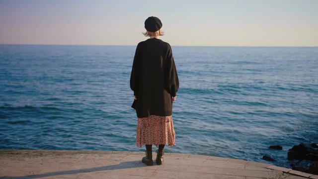 Rear view of stylish young girl in vintage outlook, dress, beret and black cardigan standing on a pier, dreamy mood shot, blue sea, waiting slow motion