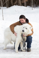 Sturdy brown-haired young man crouching on icy countryside road cuddling his huge eager Pyrenean Mountain Dog