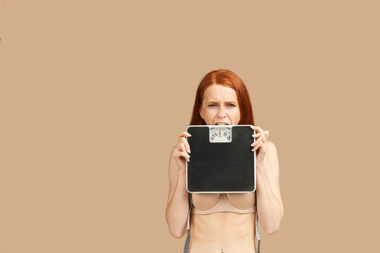 Diet and weight, young woman holding scales in hands feeling sad and depressed, trying to bite scales in dispair, isolated on beige background