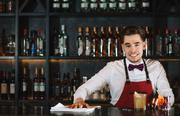 Portrait of smiling handsome young bartender, dressed in uniform with bow tie, friendly smiling at camera while standing at bar counter with shelves of bottles with alcohol on background.