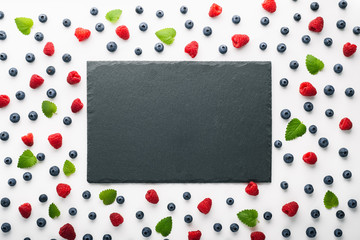 Fresh berries and mint leaves scattered around empty slate board, top view