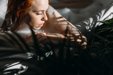 Beautiful sensual red-haired young woman relaxing in foam bathtub, enjoying sunlight and warm water in bathtub. Shadow and highlights from palm branch lying on her face, close up