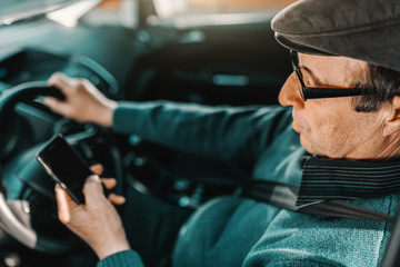 Serious Caucasian senior man with cap on head and eyeglasses sitting in car with hand on steering wheel and using smart phone. Window opened.