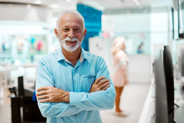 Smiling Caucasian senior man standing in tech store with arms crossed.