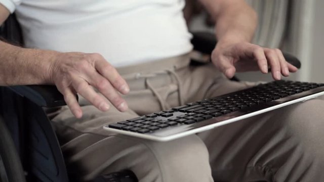 An Adult Male in a Wheelchair is Holding a Keyboard While Typing a Letter.