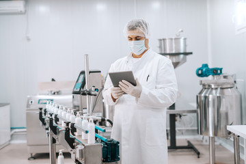 Portrait of Caucasian chemist in sterile uniform using tablet while standing next to machine with...